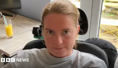 A woman with tied back brown hair wearing a grey top sits in a black padded wheelchair.