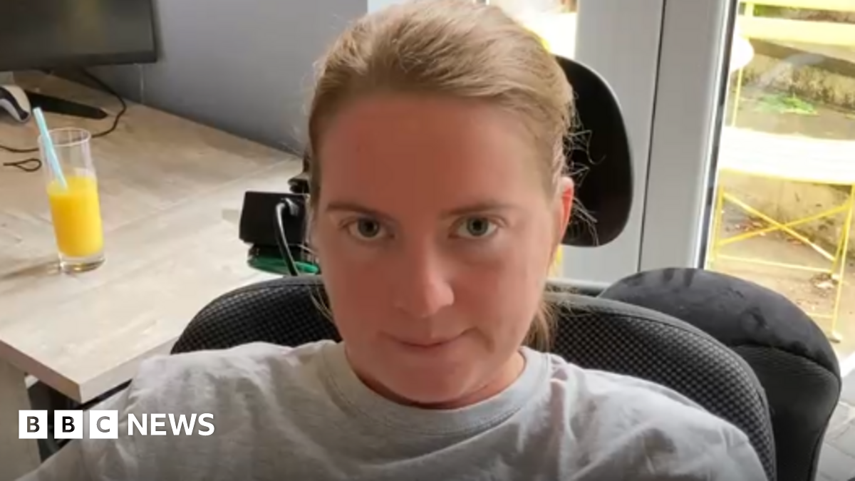 A woman with tied back brown hair wearing a grey top sits in a black padded wheelchair.