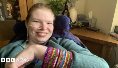 Lucinda Ritchie is smiling, seated in a supportive chair raising an arm covered with multiple colourful beaded bangles. She has an oxygen tube. She is wearing a textured, long‑sleeved top. In the background are a wooden cabinet with trophies, a desk with assorted items, and decorative plants.