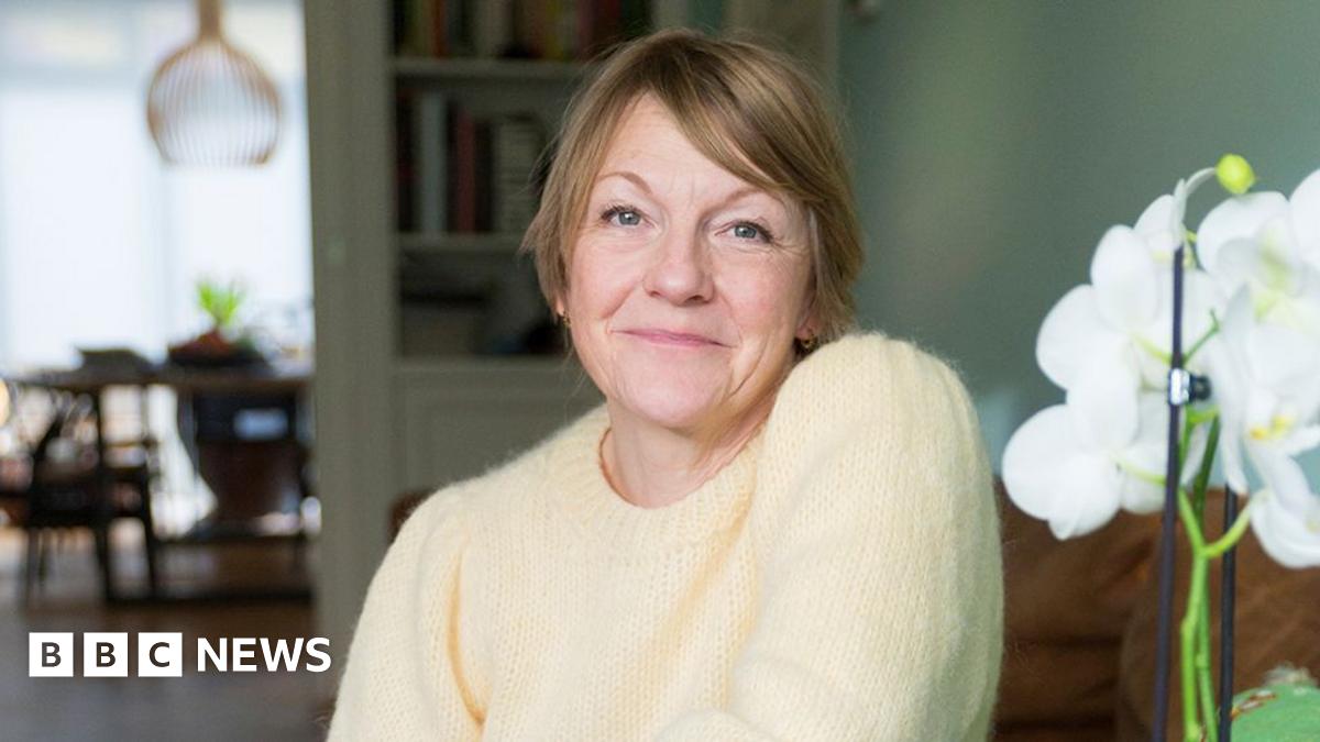 Annie sitting in a furnished room with a white flower in the foreground. She has short brown hair and is wearing a yellow jumper.