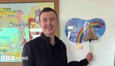 Man with dark brown short hair wears an all black outfit and is smiling - Sam Birchall. The 24-year-old stands in a hospital room in front of a bronze bell on a rainbow mount. He holds the string of the bell as if to begin ringing it.
