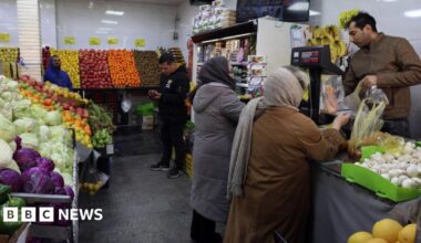 Scene inside a fruit and vegetable store, where two women are buying an item from a man at the till and two other men can be seen in the background.
