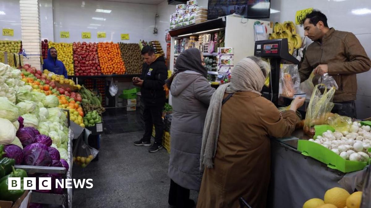 Scene inside a fruit and vegetable store, where two women are buying an item from a man at the till and two other men can be seen in the background.
