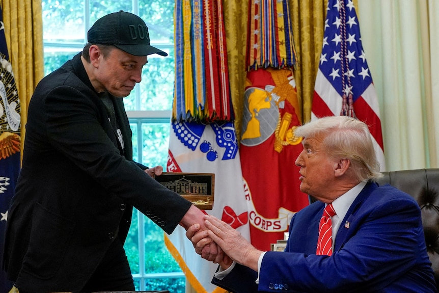 Donald Trump holds onto Elon Musk's hand with both of his own hands.