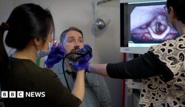 A man sits in a chair as his doctor and speech therapist view images of his vocal cords on a screen