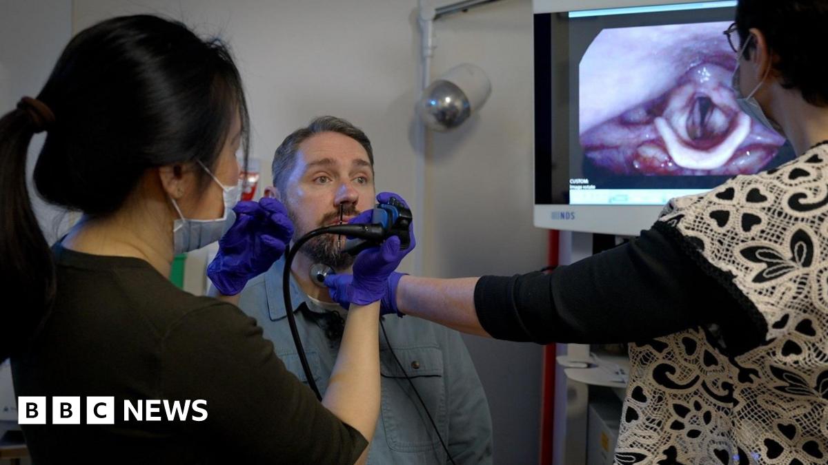 A man sits in a chair as his doctor and speech therapist view images of his vocal cords on a screen