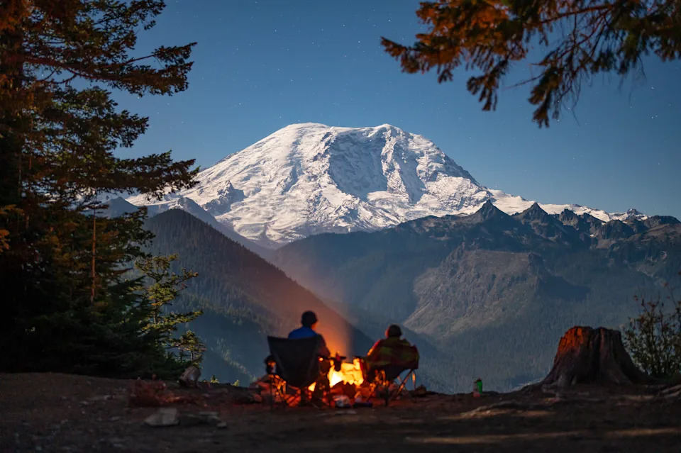 Two people sit by a campfire with a snowy mountain in the background, surrounded by trees