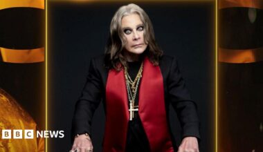 A photo of Ozzy Osbourne wearing a black jacket with large red lapels and several gold chains with crosses around his neck. He looks intensely at the camera.