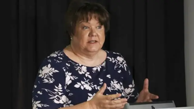 Fiona McQueen speaking behind a lectern - she is wearing a blue and white floral patterned top and has short brown hair