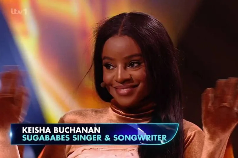 An individual, dressed in an elegant attire, is standing on a stage with hands raised, appearing to be in a moment of triumph. A name board in front of them reads "Keishna Buchanan" along with the titles "Sugababes Singer & Songwriter." The background features a vibrant and dynamic visual backdrop.