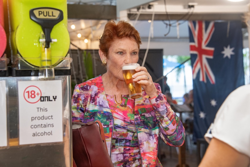Pauline Hanson sipping from a beer at the launch party. An Australian flag hanging in the background.