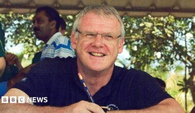 Andrew is wearing a blue polo shirt and spectacles. He in the shade, but it is a sunny day behind him. There are a few people visible in the background on the left-hand side of the image.