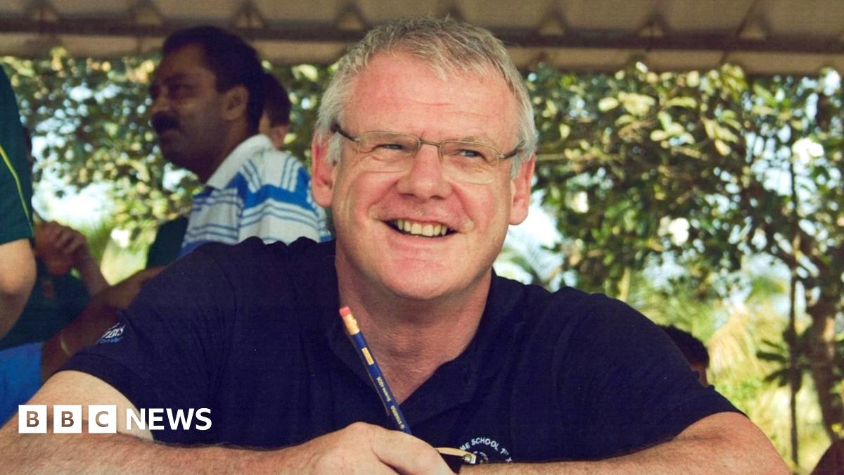 Andrew is wearing a blue polo shirt and spectacles. He in the shade, but it is a sunny day behind him. There are a few people visible in the background on the left-hand side of the image.