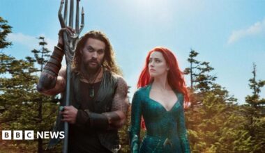 Momoa is stood in between two other people who are obscured in the image. He is wearing a pink corduroy suit and pink vest underneath and gold chain. He has his trademark long hair and beard and dark shades on.