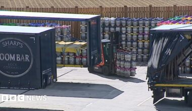 A forklift truck is carrying beer kegs across a tarmac area which has lorries and containers full of kegs. One of the containers is branded Sharp's Brewery Doom Bar.