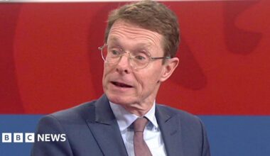 A close-up of politician Andy Street sat in a TV studio. He wears a blue suit and dark tie. His mouth is open as he is talking.