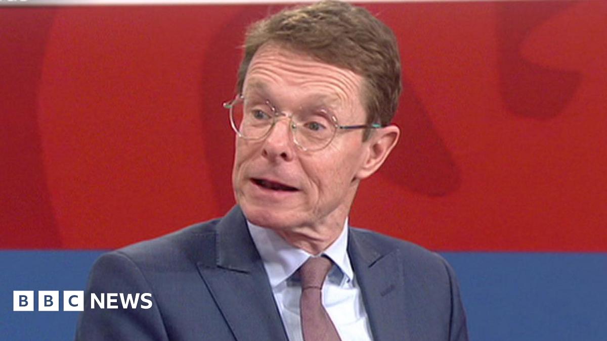 A close-up of politician Andy Street sat in a TV studio. He wears a blue suit and dark tie. His mouth is open as he is talking.