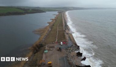 A drone view of the A379 Slapton Line between Torcross and Slapton. It shows the sea on one side and a lake on the other side. The road is damaged in two parts where maintenance vans can be seen.