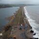 A drone view of the A379 Slapton Line between Torcross and Slapton. It shows the sea on one side and a lake on the other side. The road is damaged in two parts where maintenance vans can be seen.