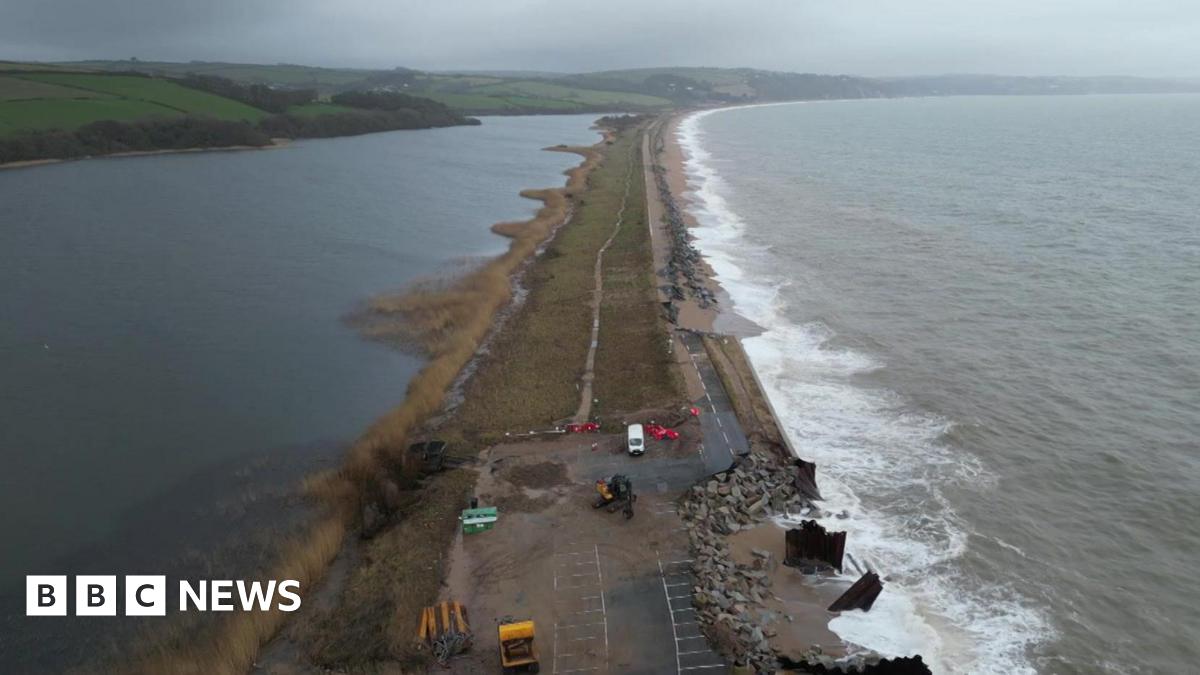 A drone view of the A379 Slapton Line between Torcross and Slapton. It shows the sea on one side and a lake on the other side. The road is damaged in two parts where maintenance vans can be seen.