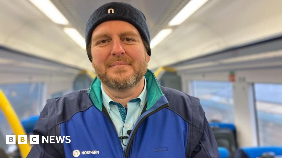 Paul is wearing a Northern uniform, standing in a railway carriage, smiling at the camera.