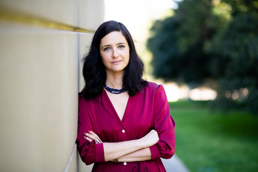 A young white woman with long dark hair leaning against a building