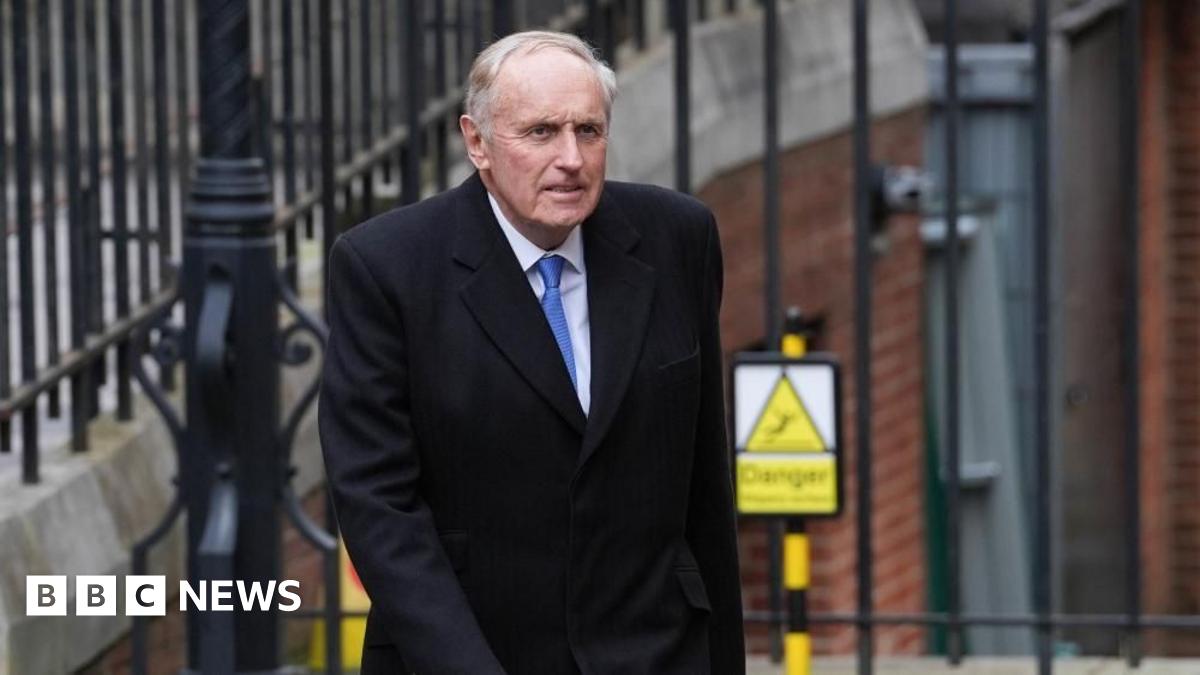 Dacre walking in front of a black metal fence. He wears a long black coat, a white shirt and blue tie. He looks sincere