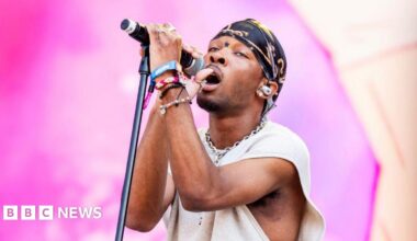 D4vd holds a microphone stand close to him while performing onstage during a music festival in 2024. He has a black and gold headscarf on and a cream top.