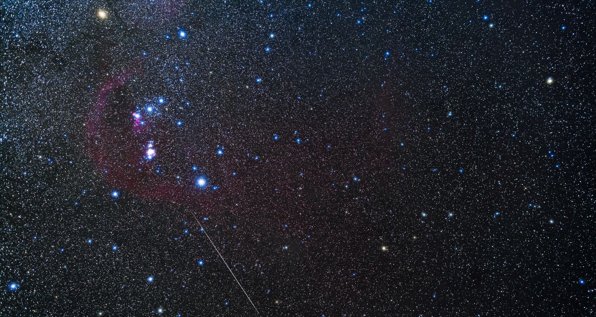 The stars of the constellation Orion shine in a winter night sky against a background of fainter stars as a meteor streaks diagonally across the image to the lower right.