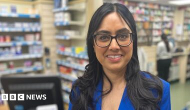 Jasvinder Kaur Lali a Superintendent Pharmacist standing at the chemist shop counter of the Carlton Hill Pharmacy in Nottinghamshire. She is wearing a blue jacket and white blouse