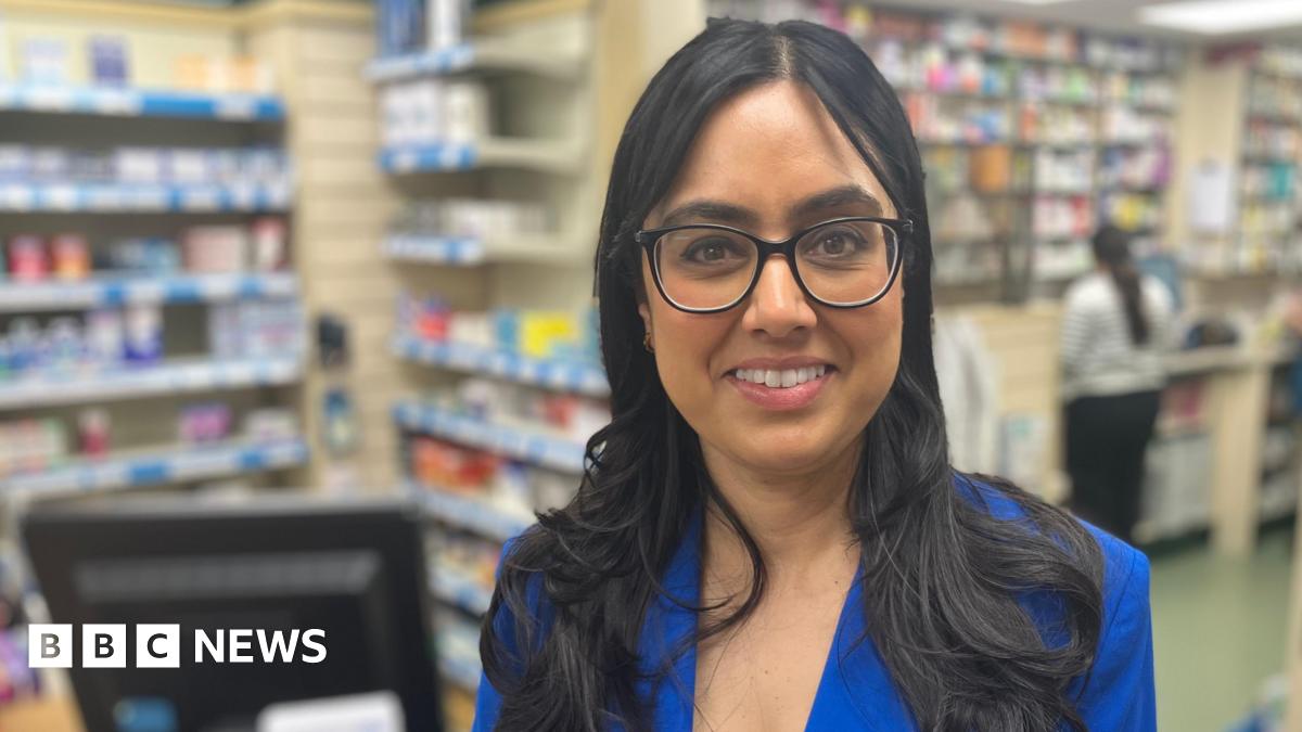 Jasvinder Kaur Lali a Superintendent Pharmacist standing at the chemist shop counter of the Carlton Hill Pharmacy in Nottinghamshire. She is wearing a blue jacket and white blouse