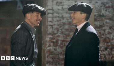 Two actors in formal dress wearing flat caps in conversation. One man is Cillian Murphy stood on the right, the other is Barry Keoghan.