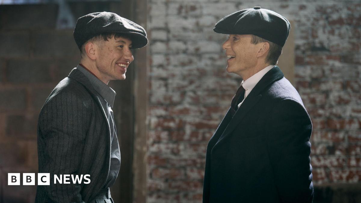 Two actors in formal dress wearing flat caps in conversation. One man is Cillian Murphy stood on the right, the other is Barry Keoghan.