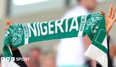 A Nigeria football fan holds a green T-shirt with the words 'Hope Dey' (meaning 'there is hope') written in white and 'we must qualify' underneath. He is surrounded by other Nigeria fans who are seated in a stadium