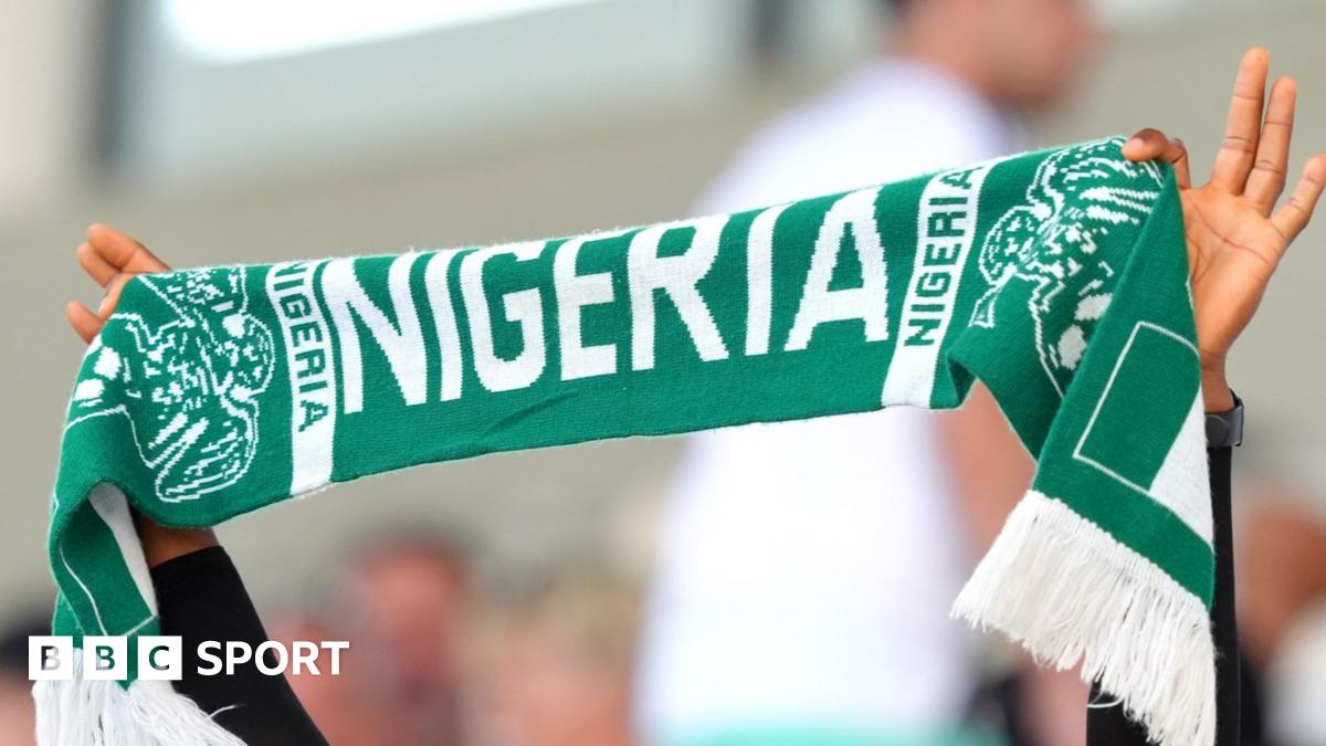 A Nigeria football fan holds a green T-shirt with the words 'Hope Dey' (meaning 'there is hope') written in white and 'we must qualify' underneath. He is surrounded by other Nigeria fans who are seated in a stadium
