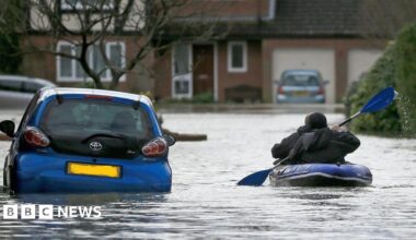 A resident uses a boat to make their way through floodwater in Chertsey in 2014. Residential houses can be seen in the background, while cars are also surrounded by water.
