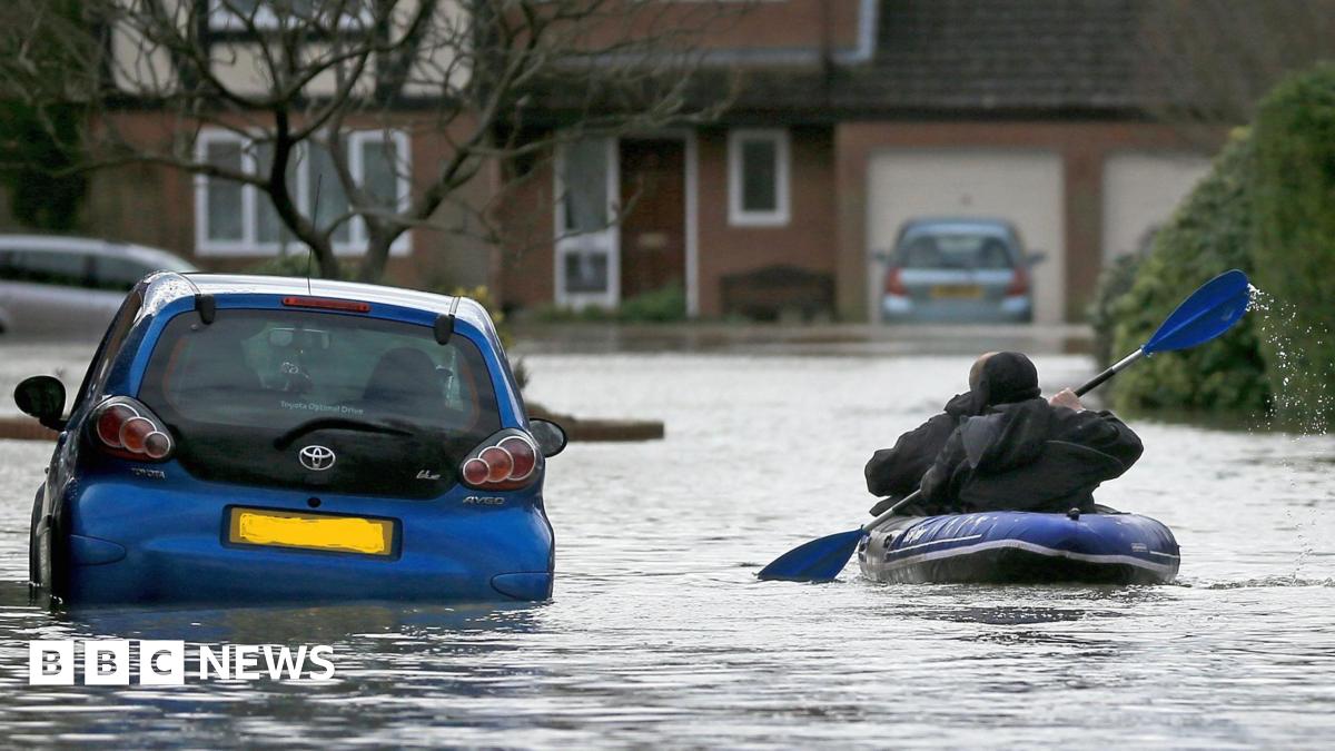 A resident uses a boat to make their way through floodwater in Chertsey in 2014. Residential houses can be seen in the background, while cars are also surrounded by water.