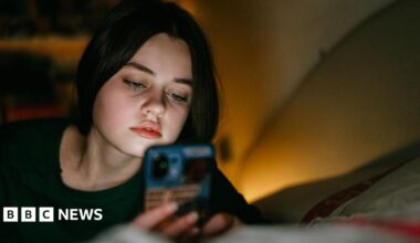 A woman looks at her phone lying on the bed in a low light.