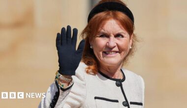 Sarah Ferguson waves her black-gloved hand while wearing a black headband, a cream jacket with a black trim, and large pearl earrings.