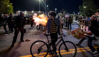 Man standing with bicycle as riot police push protesters back behind him
