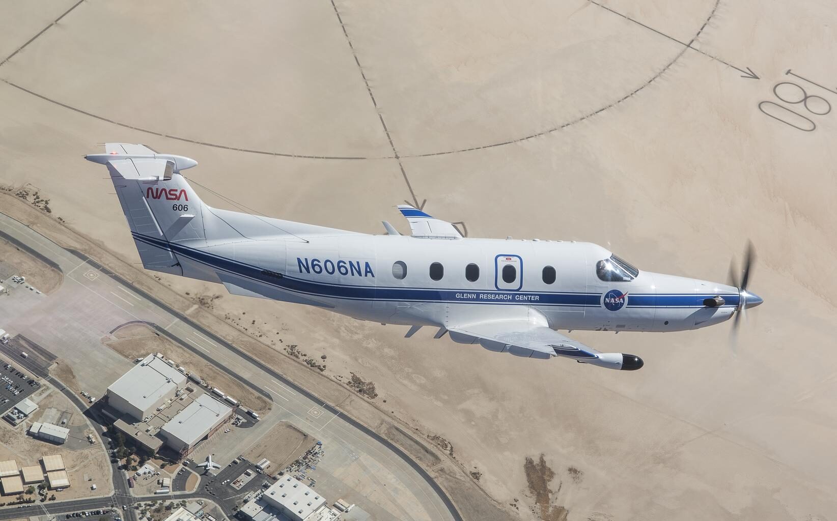 NASA’s freshly painted Pilatus PC-12 flies over NASA’s Armstrong Flight Research Center in Edwards, California. The parking lots and center buildings dot the landscape below. The compass rose in the upper part of the photo shaped like the sun hovers over the aircraft, emphasizing the gleaming white plane with a blue stripe and blue N606NA number across the fuselage and NASA red worm logo on the tail. A sensor, with a black-tip juts out from below the wing.