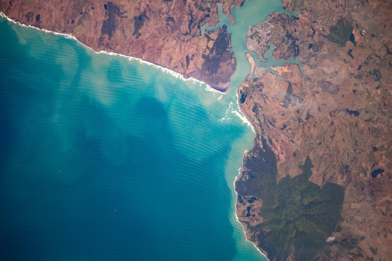 Aerial view of a coastal landscape showing turquoise ocean waves meeting a rugged, brown shoreline with a river inlet, small town, and patches of green vegetation inland.