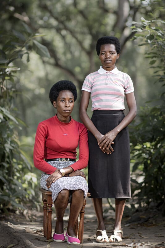 Two women pose for a portrait outdoors; one sits on a small wooden chair wearing a red top and patterned skirt, while the other stands beside her in a striped polo shirt and dark skirt. Green foliage surrounds them.