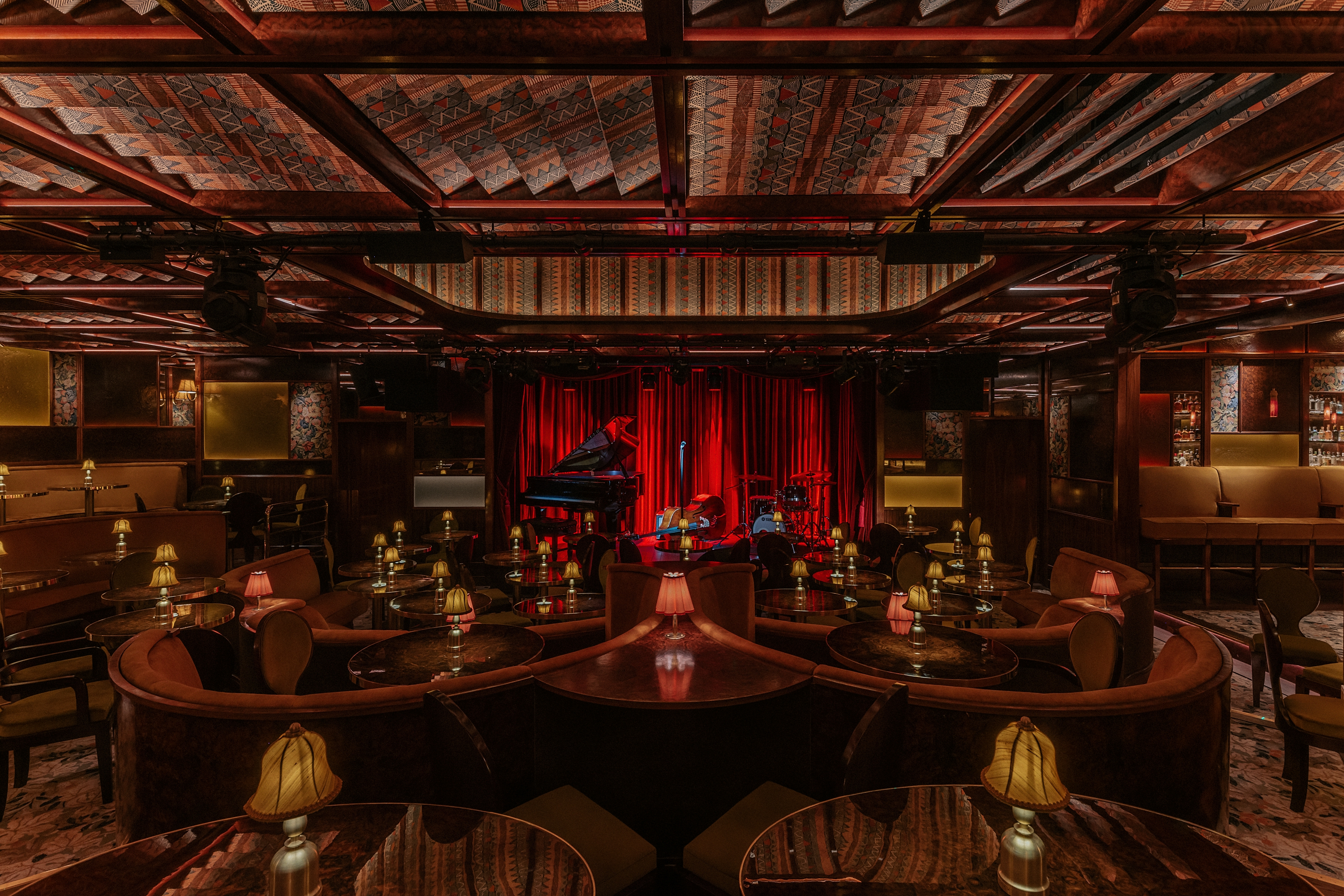 A dimly lit, textiles and lacquered wood-clad jazz club with lots of softly glowing table lamps, performers playing live, and a red-tinted light.