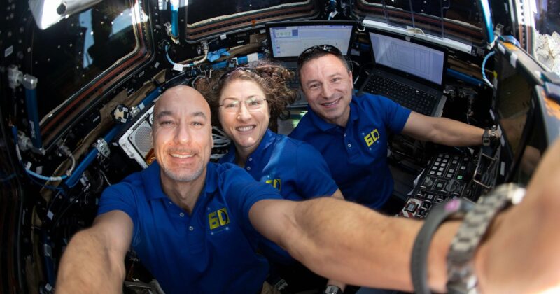 Three astronauts in blue shirts smile and pose for a selfie inside a spacecraft, surrounded by control panels, screens, and technical equipment. Bright light comes through the windows behind them.