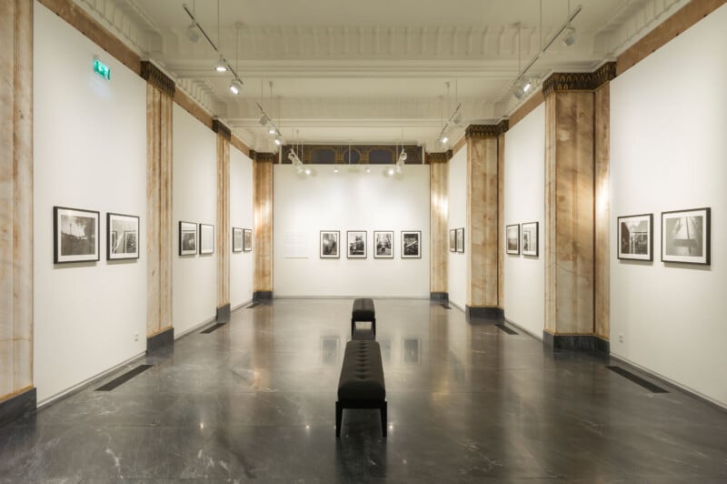 A spacious, elegant art gallery with marble columns, gray marble floor, white walls, and black cushioned benches. Framed black-and-white photographs are evenly displayed on the walls under bright ceiling lights.