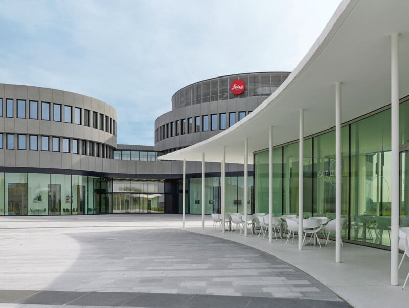 A modern building complex with curved glass walls, white pillars, and outdoor seating. The Leica logo is visible on a round, grey building in the background under a clear sky.