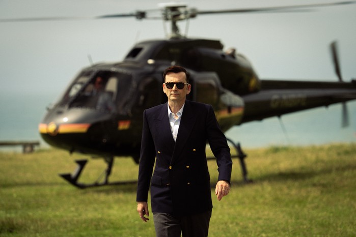 David Tennant as Lord Tony Baddingham striding across a field in a double-breasted blazer and sunglasses, walking away from his black helicopter.