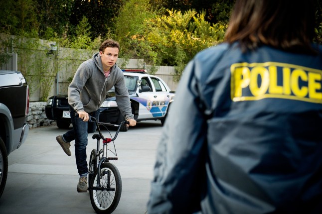 Finn Cole in a scene from Animal Kingdom, riding a bike while looking at a female police officer whose face is not seen.