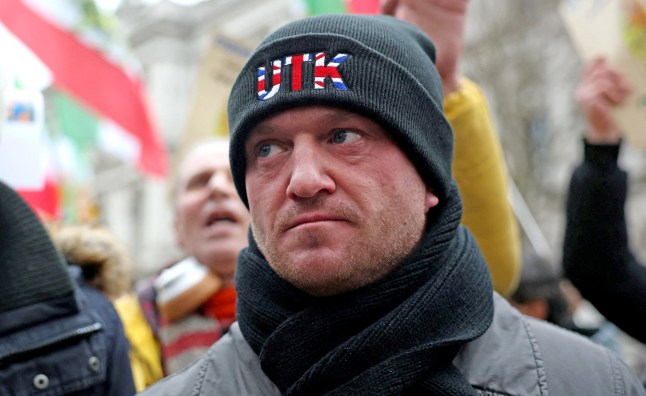 A close-up of Tommy Robinson (Stephen Yaxley-Lennon) at a protest wearing a black scarf and black hat, which reads 'UTK' in a red, white and blue pattern.